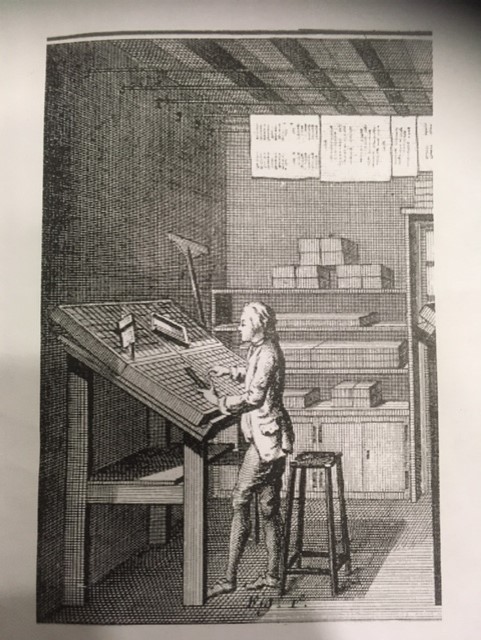 man standing at his wooden desk choosing a type from his case. In the background there is paper hanging from the ceiling.